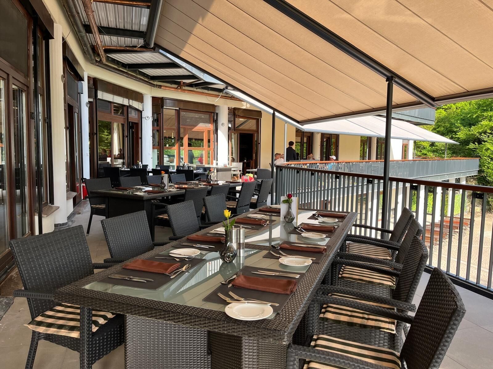 An outdoor dining area with glass-top tables and wicker chairs, surrounded by a covered patio with a view of a building and greenery.