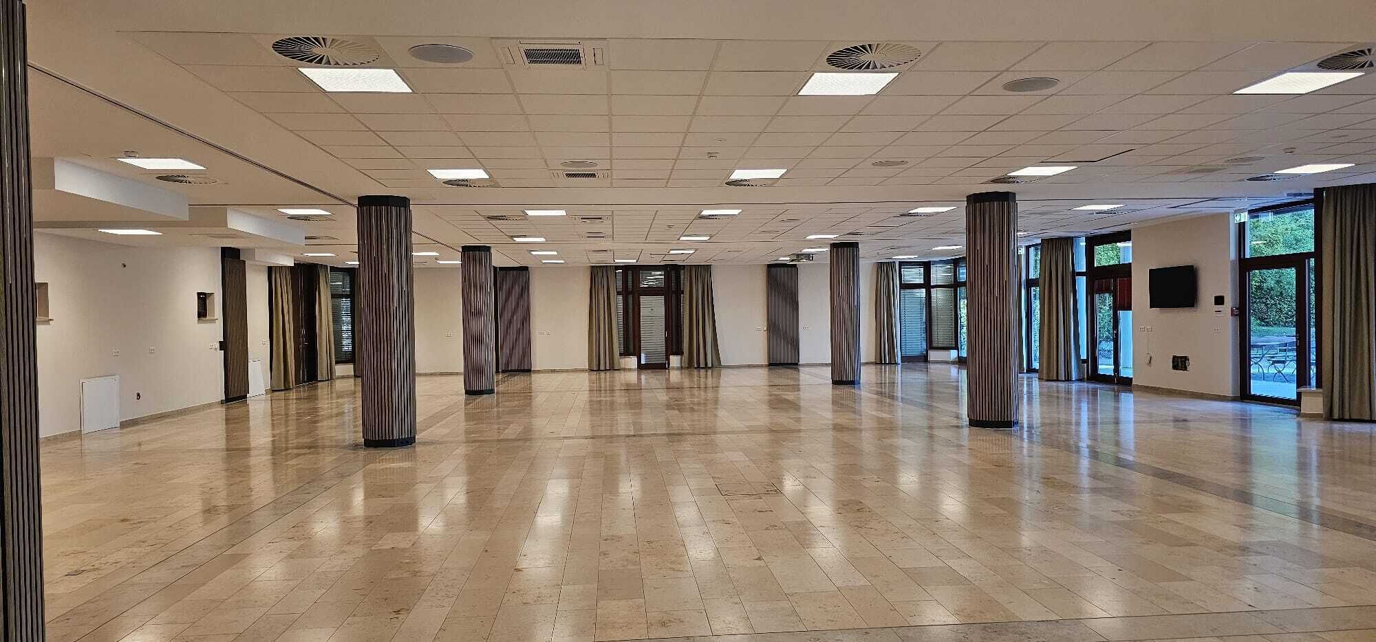 Un amplio y luminoso vestíbulo de hotel, con suelo de madera pulida, altas columnas y grandes ventanales que permiten la entrada de luz natural.