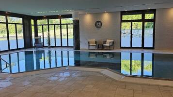 An indoor swimming pool with clear glass walls, a clock on the wall, and a seating area with chairs and a table.