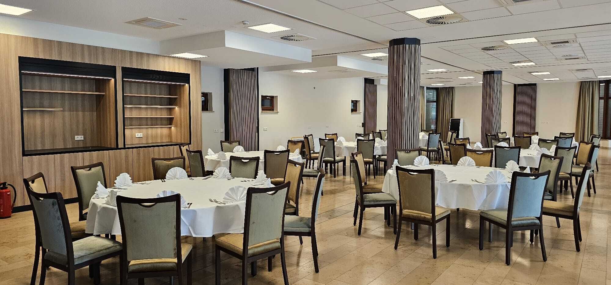 Uma sala de banquetes com várias mesas redondas preparadas para refeições, cada uma com toalhas brancas e cadeiras.