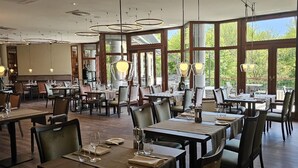 Un restaurante espacioso con techos altos, amplias ventanas e iluminación colgante moderna. Las mesas están ordenadas con sillas, y el interior combina elementos de madera y metal.