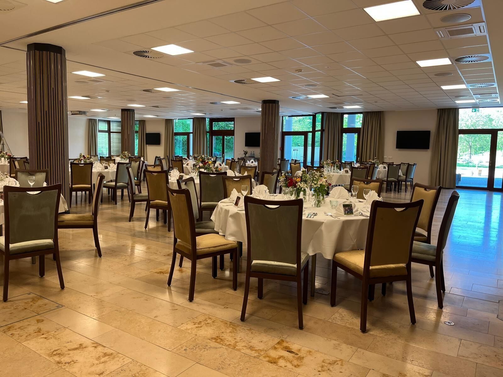 Un amplio salón de banquetes con varias mesas redondas dispuestas para comer, ventanas grandes que ofrecen luz natural y un techo alto con iluminación empotrada.