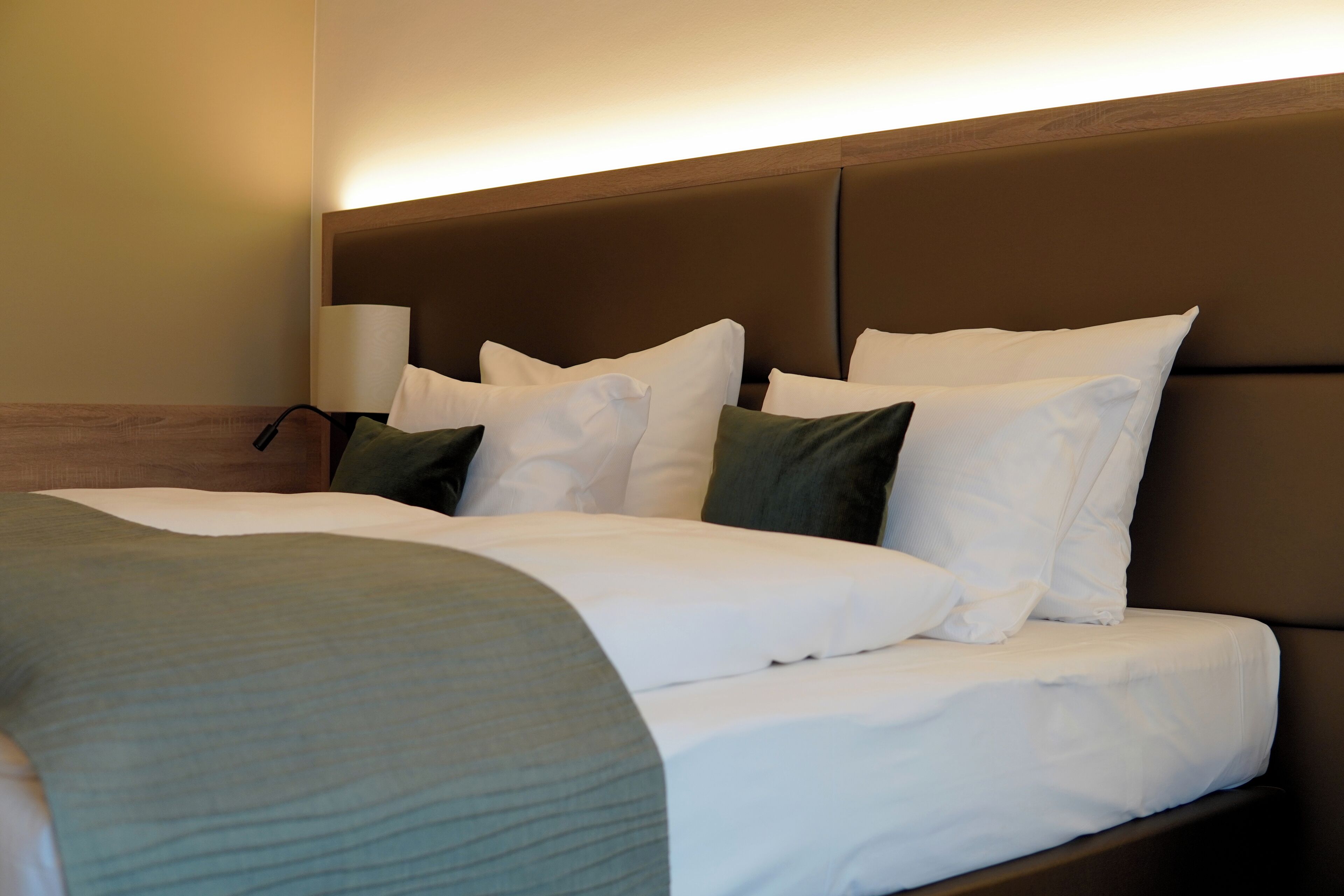 Cama bem arrumada com roupa de cama branca e verde, cabeceira de madeira e abajur.