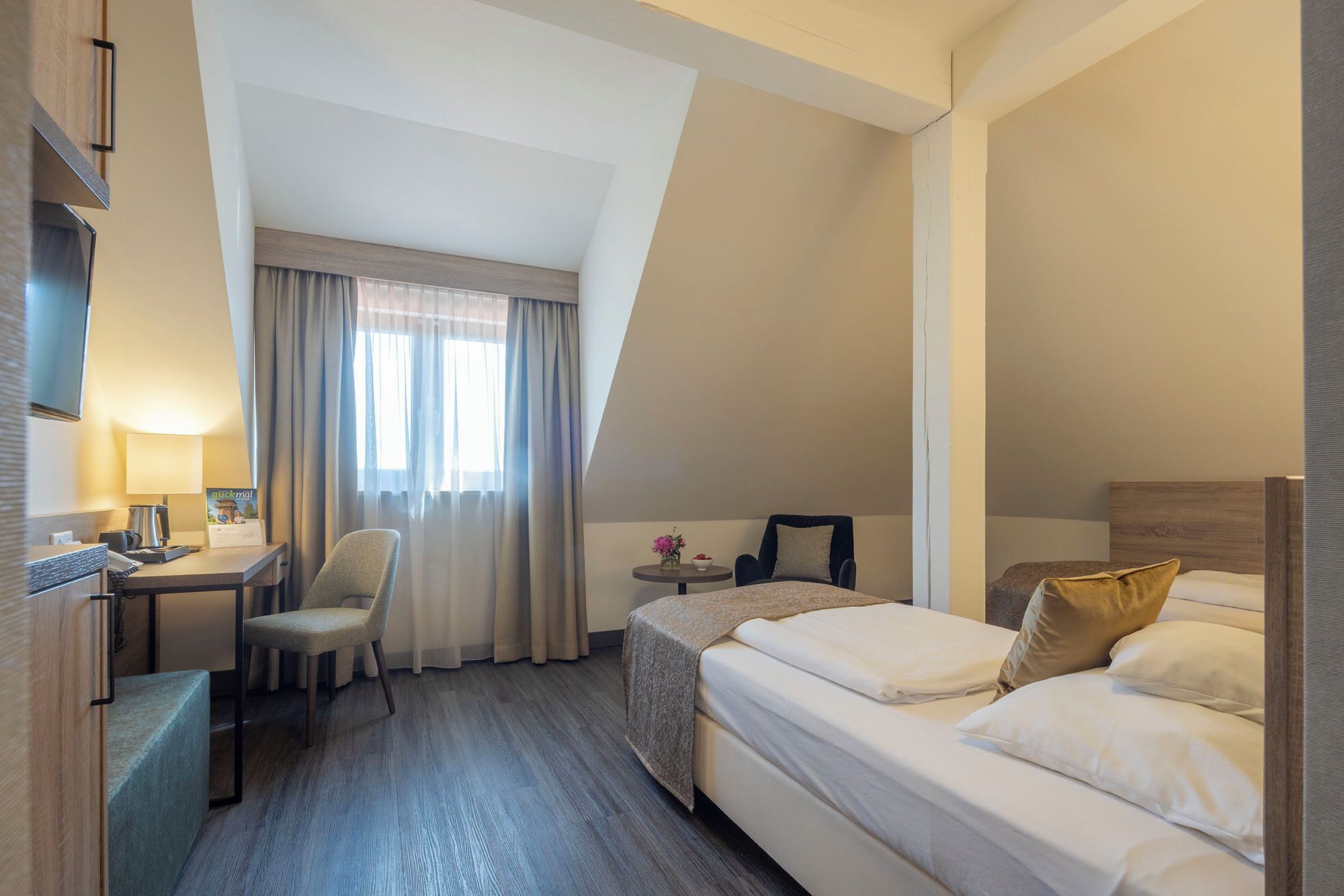 A hotel room with a bed, a desk, a chair, a small table with a vase, and a window with curtains.
