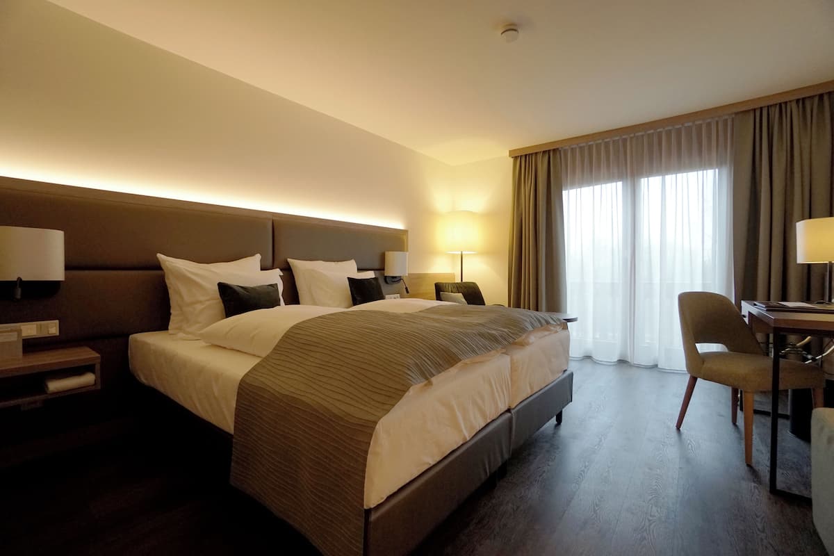 A hotel room with a large bed, two bedside tables with lamps, a desk with a chair, and a large window with curtains.