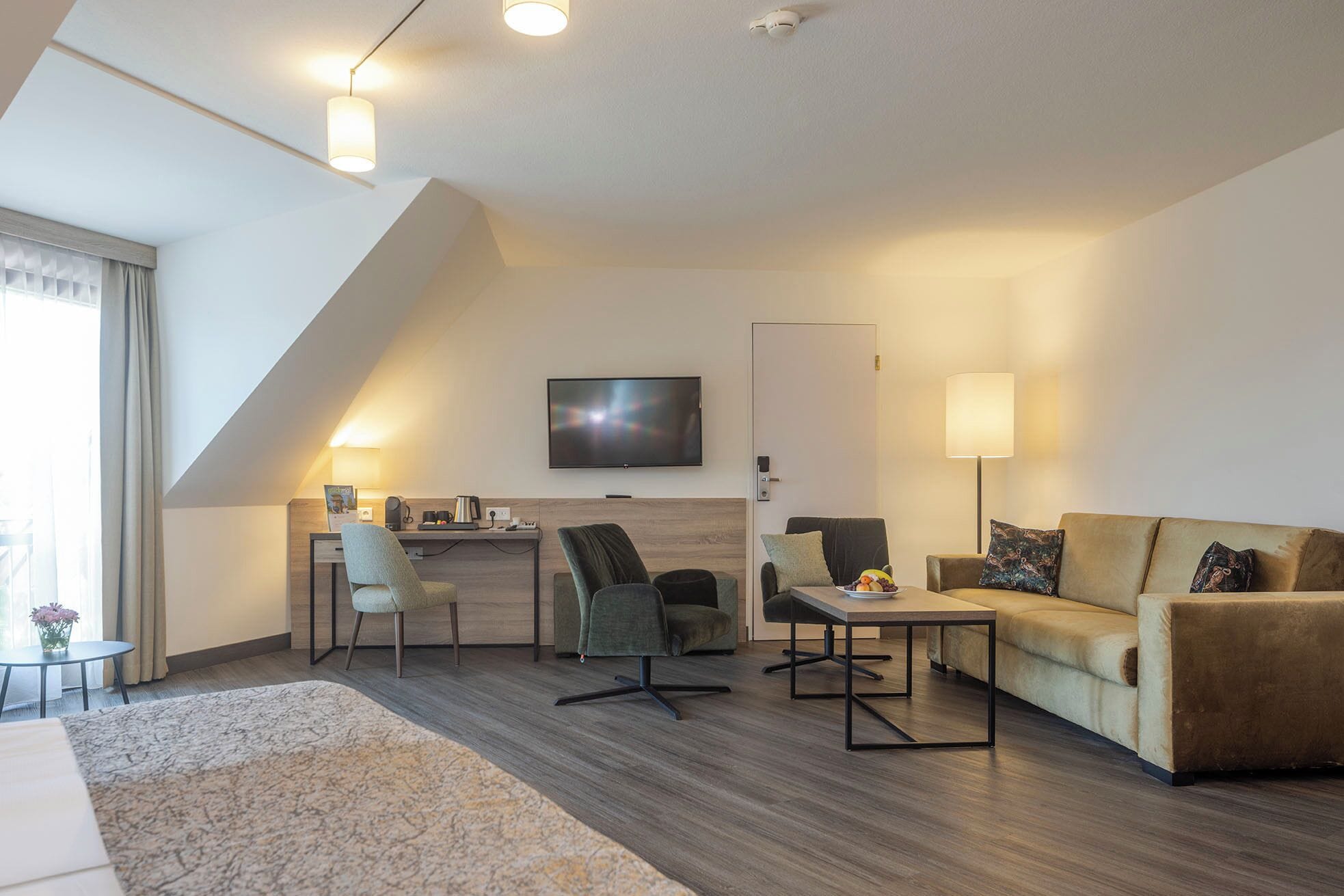 Un salon moderne comprenant un canapé, deux fauteuils, une table basse et un bureau avec un ordinateur. Un téléviseur à écran plat est fixé au mur.