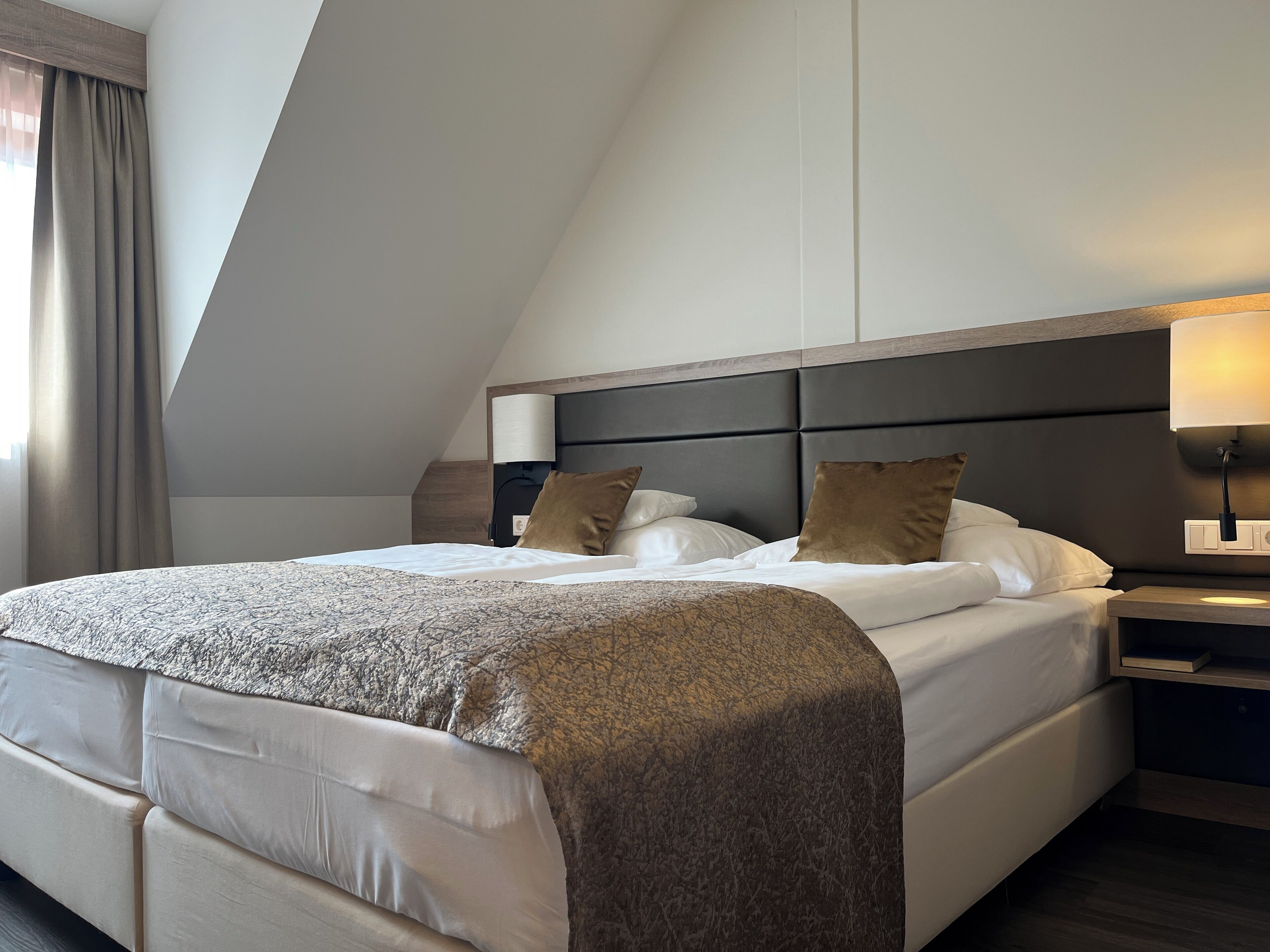 Ein modernes Hotelzimmer mit einem großen Bett, einem Nachttisch, einer Lampe und Blick auf die Stadt durch das Fenster.