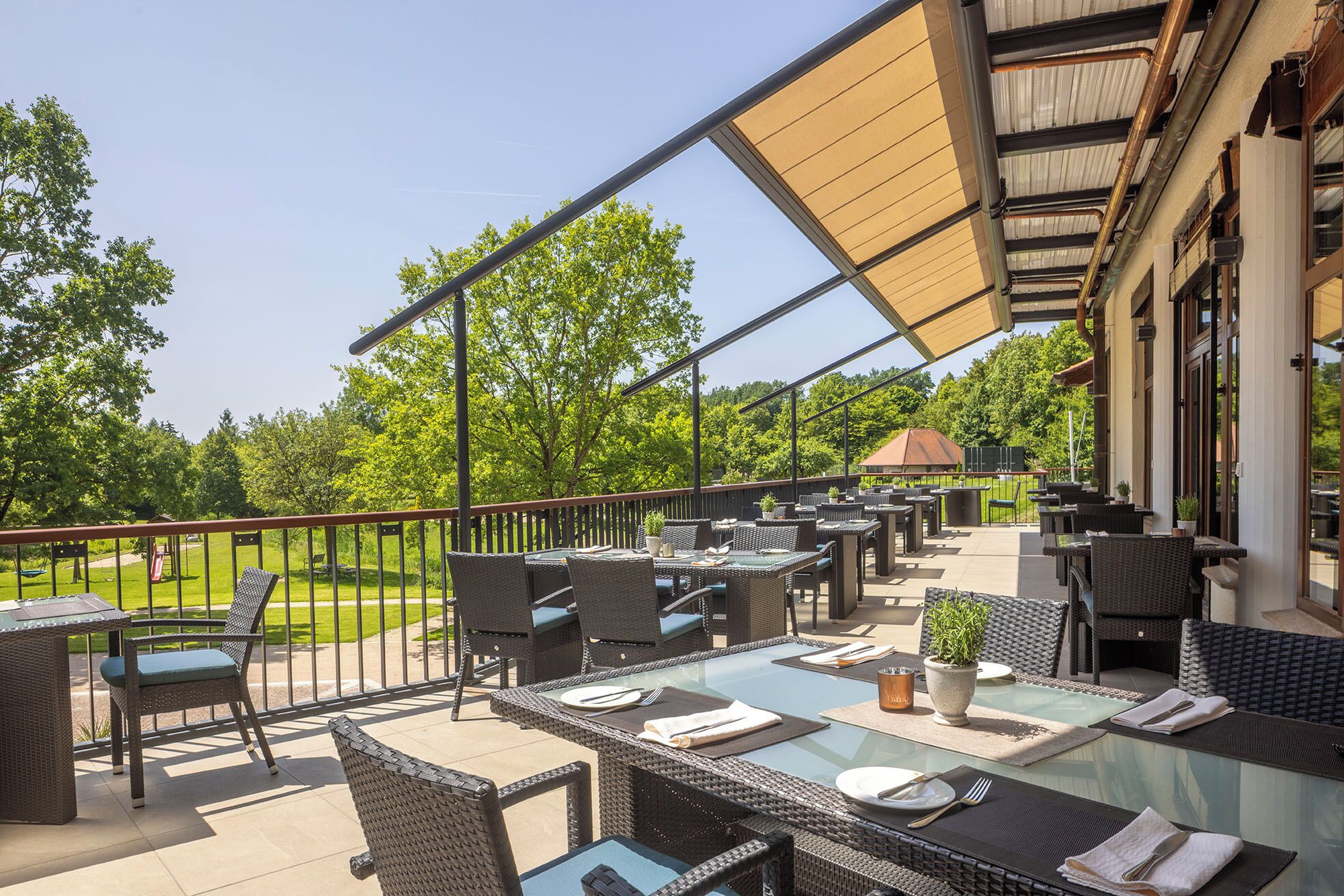 An outdoor patio with wicker furniture, glass tables, and a view of a green park.
