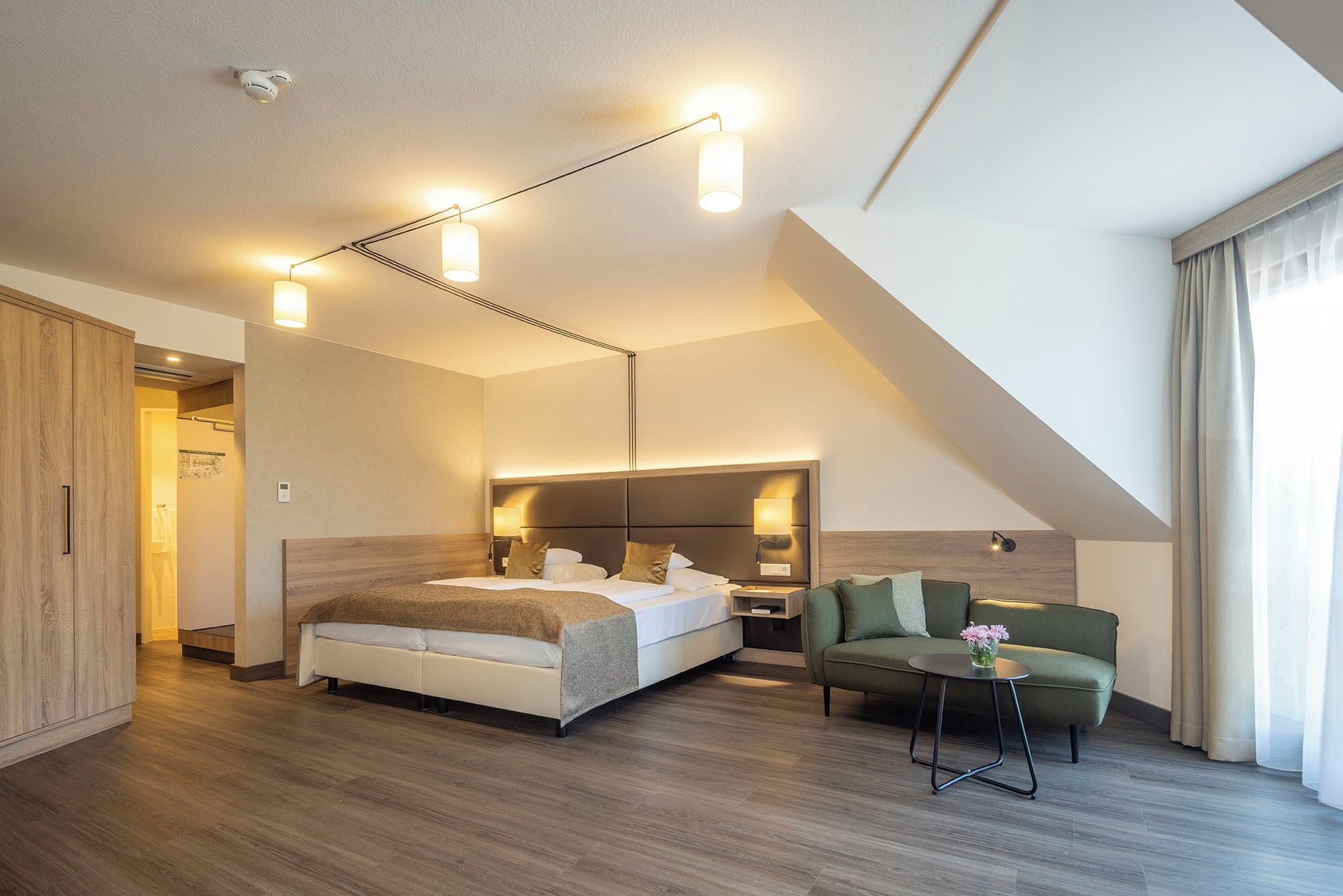 Ein modernes Hotelzimmer mit einem großen Bett, einer Couch und einem kleinen Tisch mit Blumen. Deckenleuchten und ein Fenster mit Vorhängen sind vorhanden.