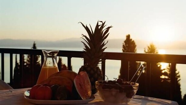 A table with a fruit bowl, a glass of juice, and a pineapple on a balcony overlooking a sunset. - Aronis Apartments (Corfu)