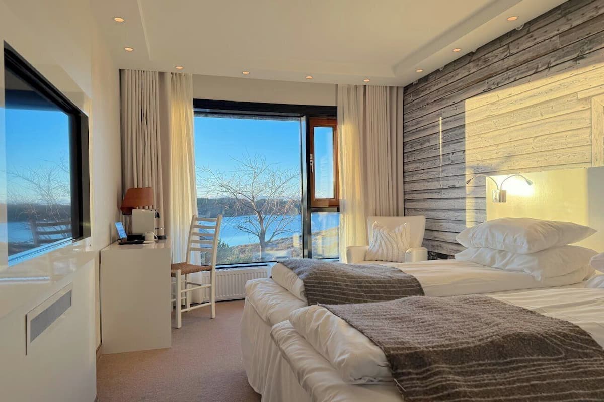 A modern hotel room with two beds, a desk, and a view of the outdoors.
