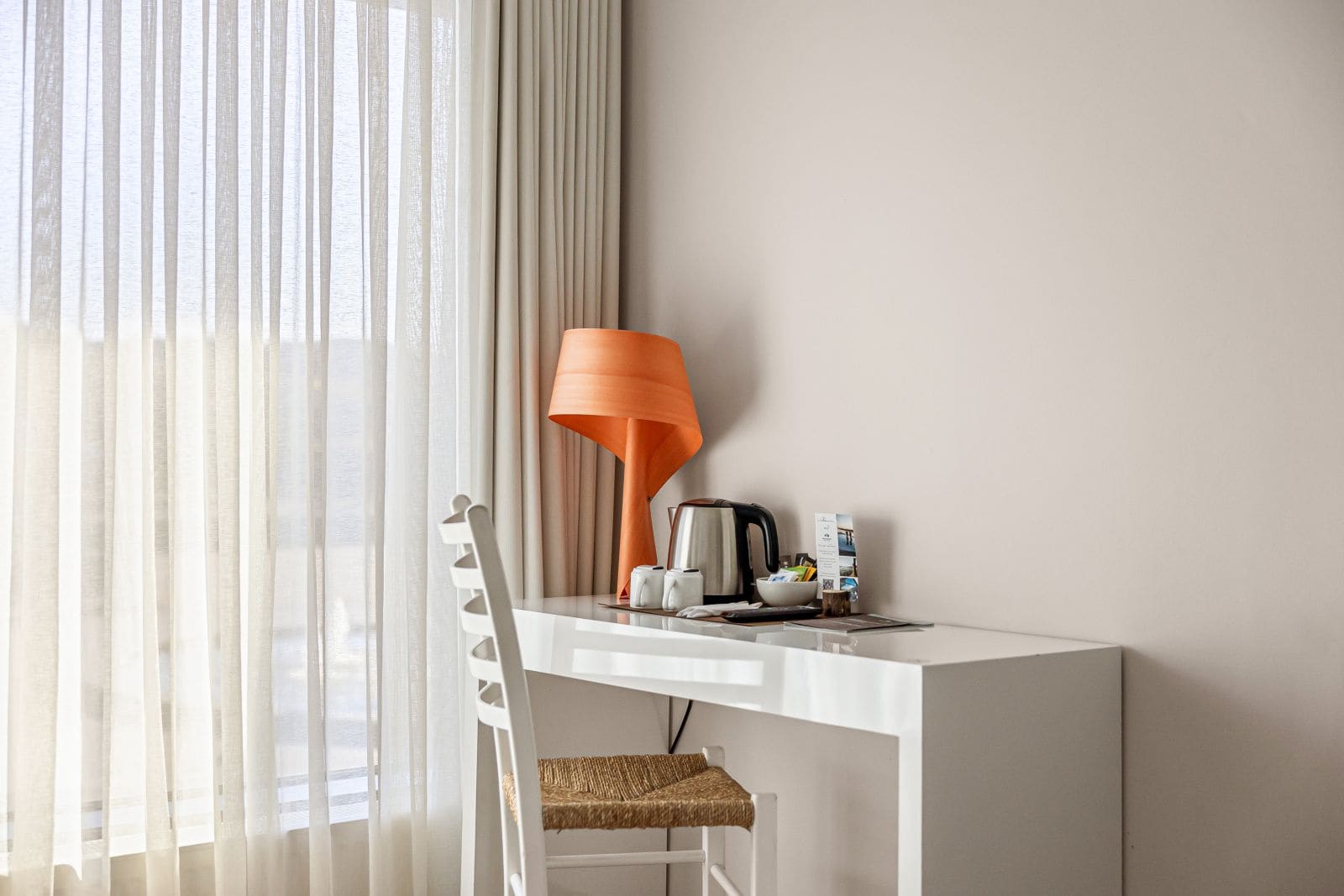 A hotel room with a white desk, a wicker chair, a white lamp, a kettle, and a small table with a cup and a brochure.