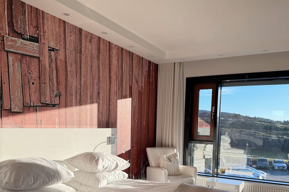 A modern bedroom with a large bed, a chair, and a view of a parking area through a large window.