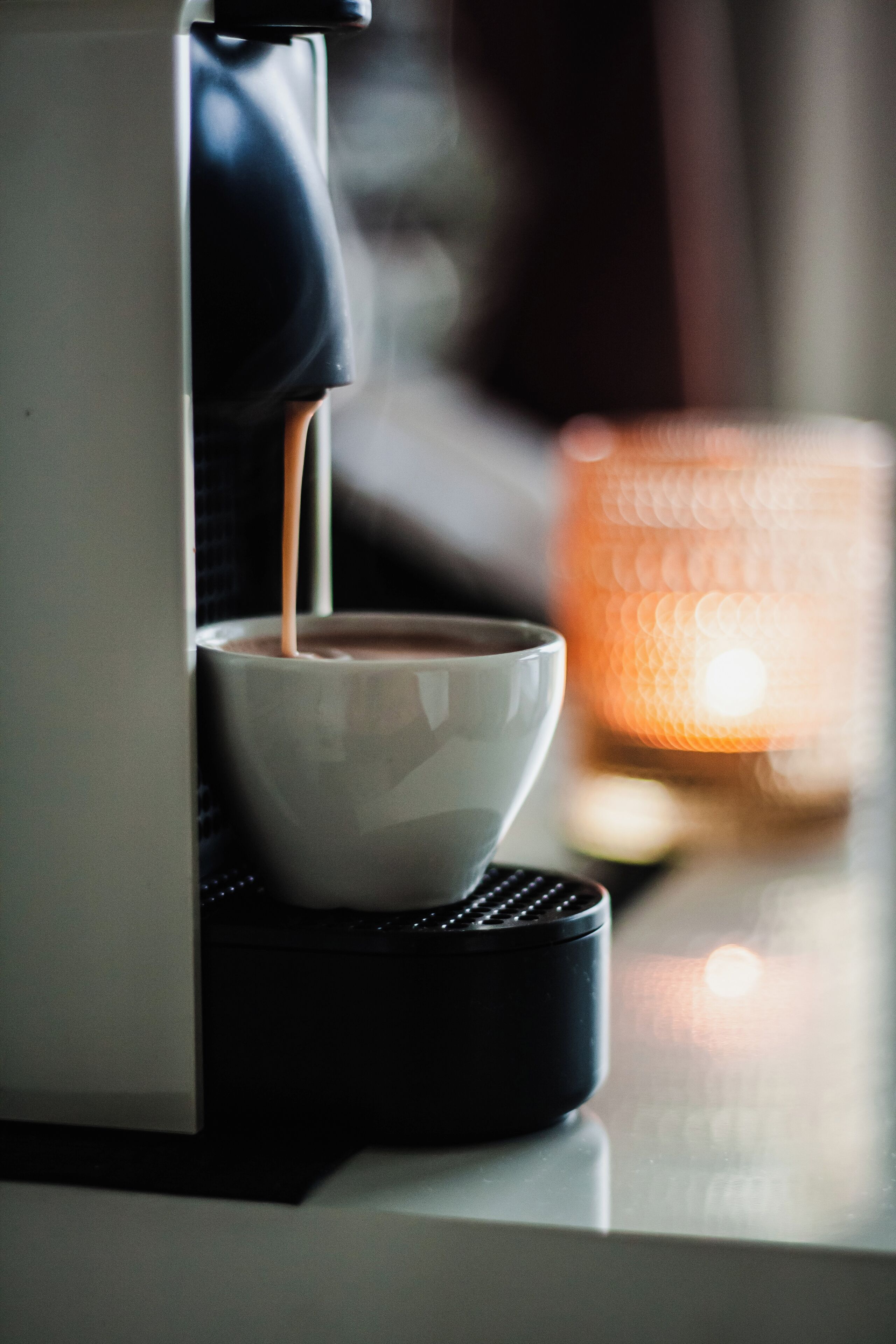 Una cafetera sirviendo café en una taza blanca sobre un posavasos negro, con una vela encendida al fondo.