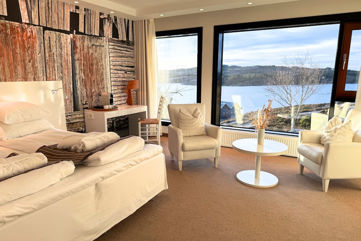 A modern bedroom with a large bed, two white armchairs, a round coffee table, and a view of a lake and hills.