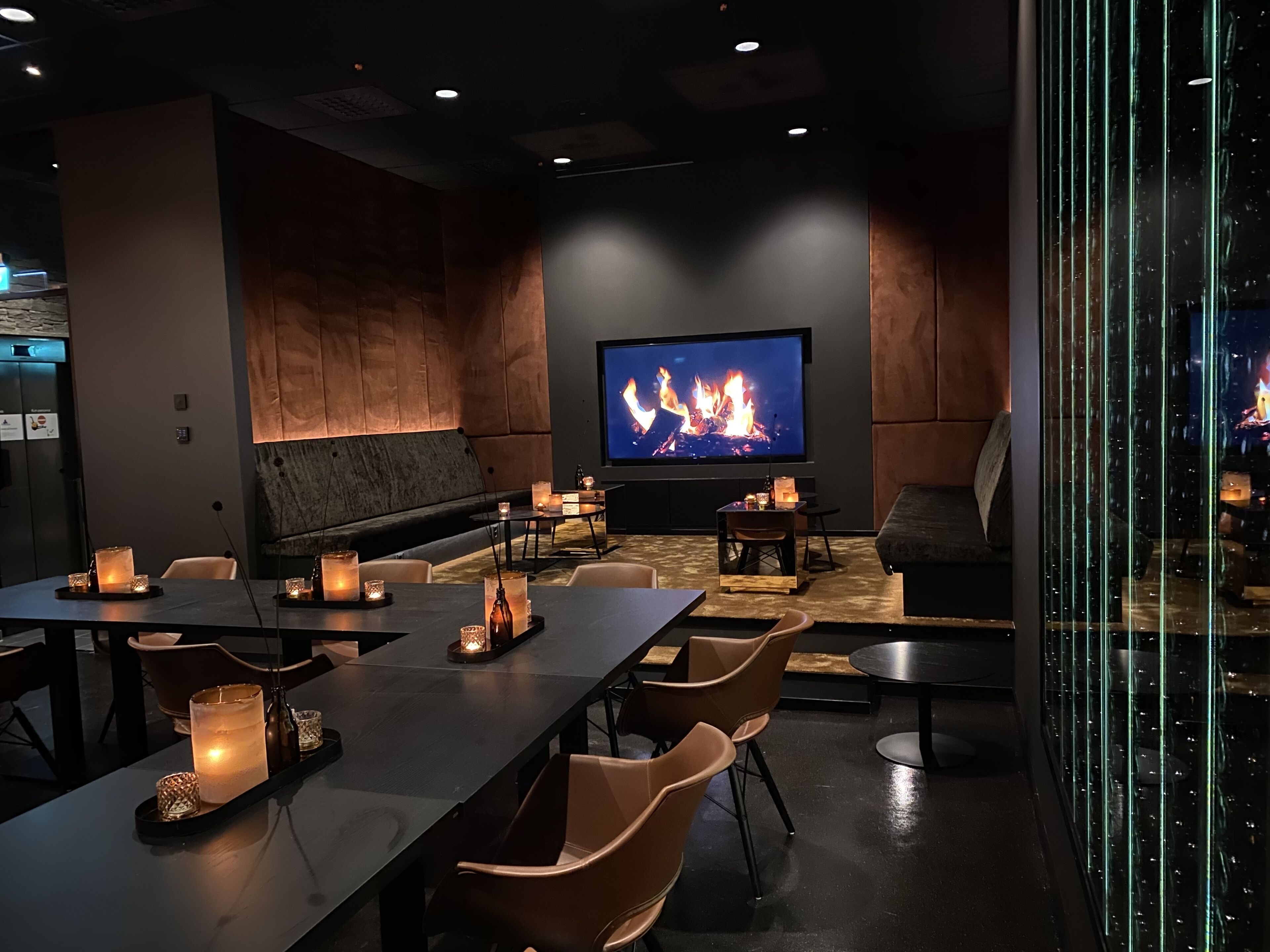 A modern lounge area with a large flat-screen TV displaying a fire, comfortable seating, and tables with candles.