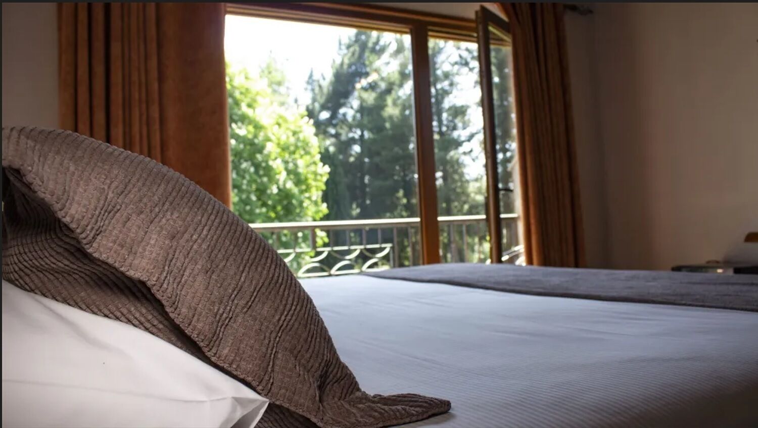 A bed with a white pillow and a brown blanket, a window with curtains, and a view of trees outside.