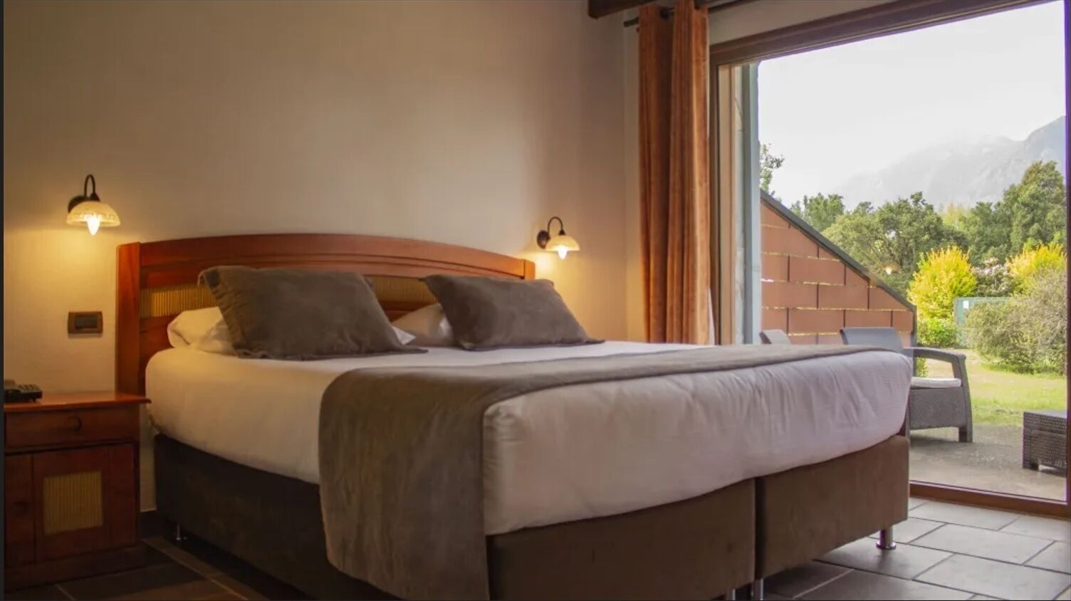 A bed with a headboard, two bedside lamps, and a view of a balcony with outdoor furniture.