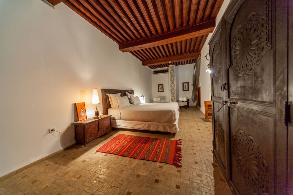 A bedroom with a wooden ceiling, a bed, a nightstand, a lamp, a wardrobe, and a patterned rug.