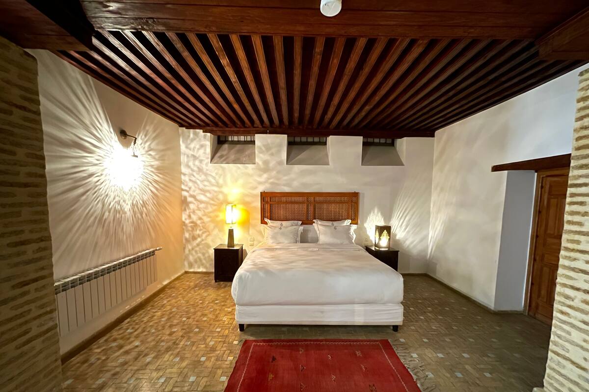 A bedroom with a large bed, wooden ceiling beams, a red rug, and two bedside lamps.