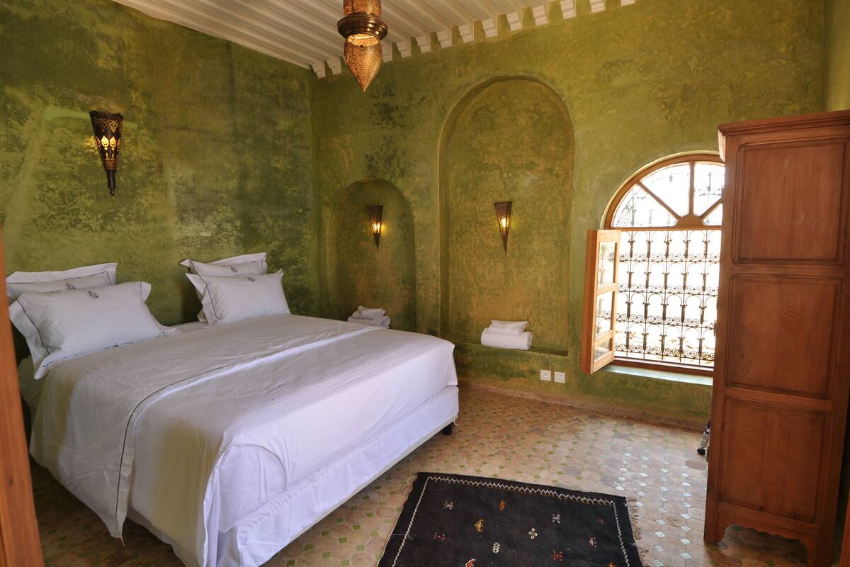 A bedroom with a large bed, two wall-mounted lights, a wooden wardrobe, and a window with a metal gate.