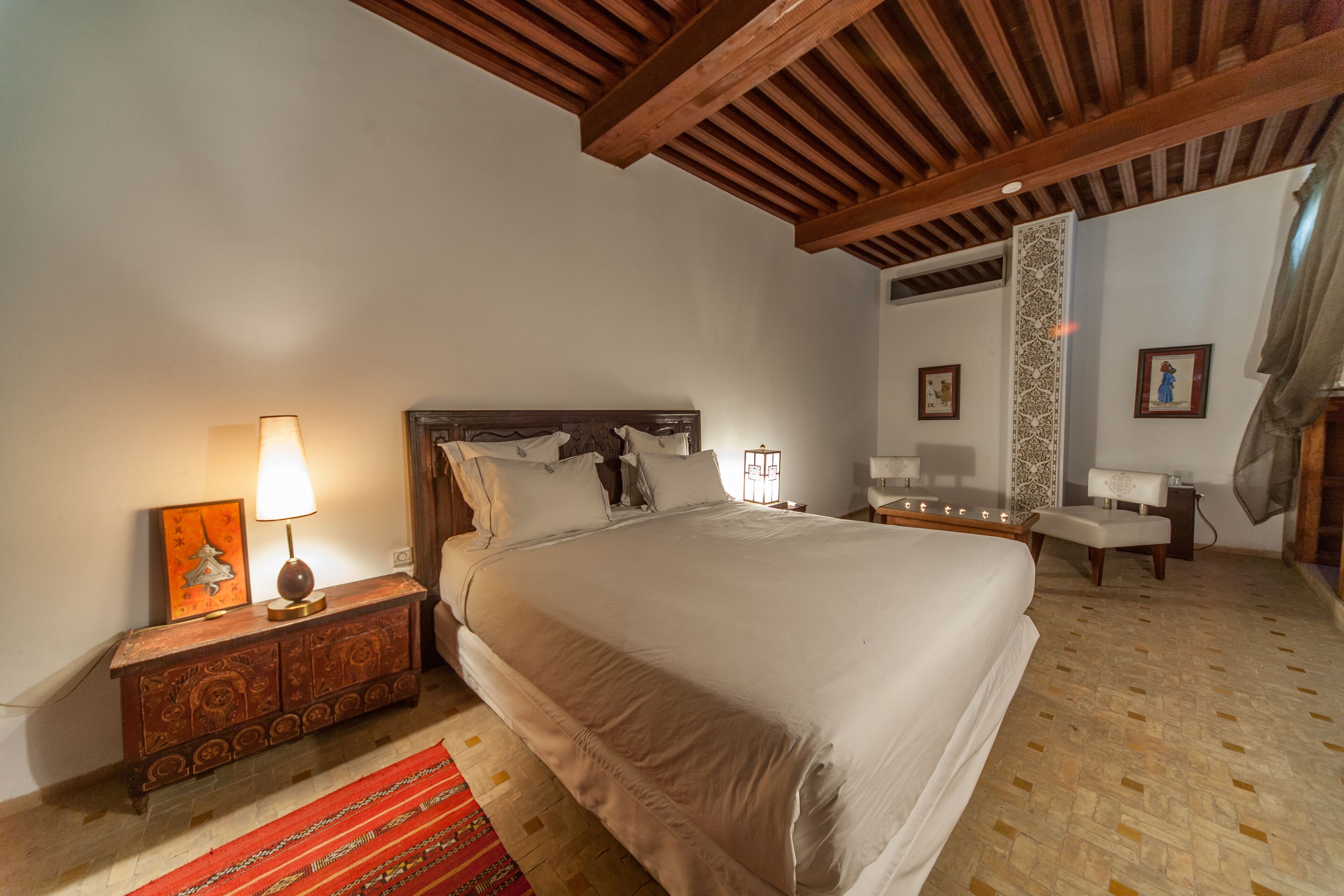 A bedroom with a large bed, wooden headboard, bedside tables with lamps, a red patterned rug, and a decorative wall panel.