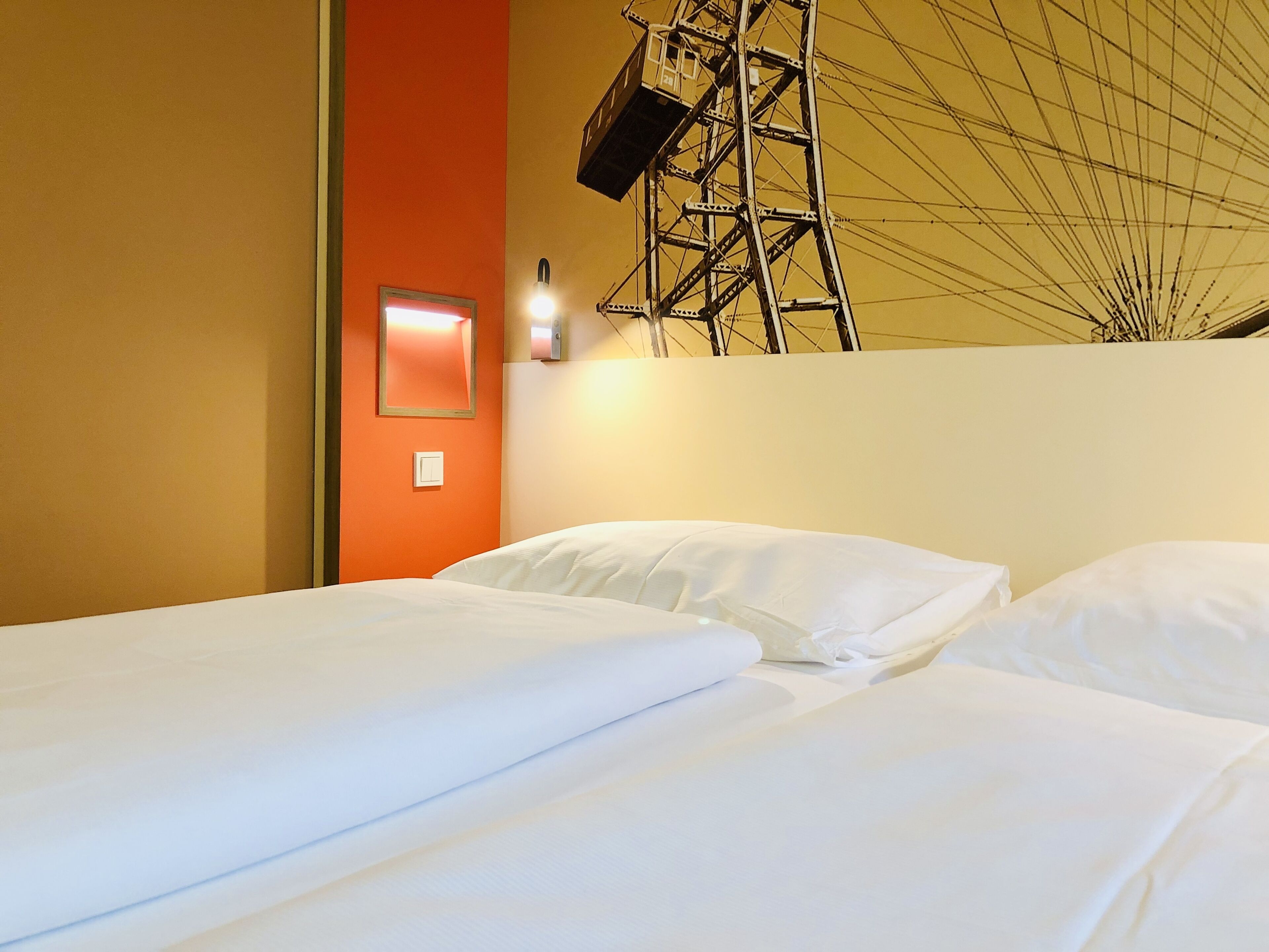 Une chambre d’hôtel avec deux lits, un mur d’accent rouge et une grande fenêtre donnant sur un bâtiment imposant.