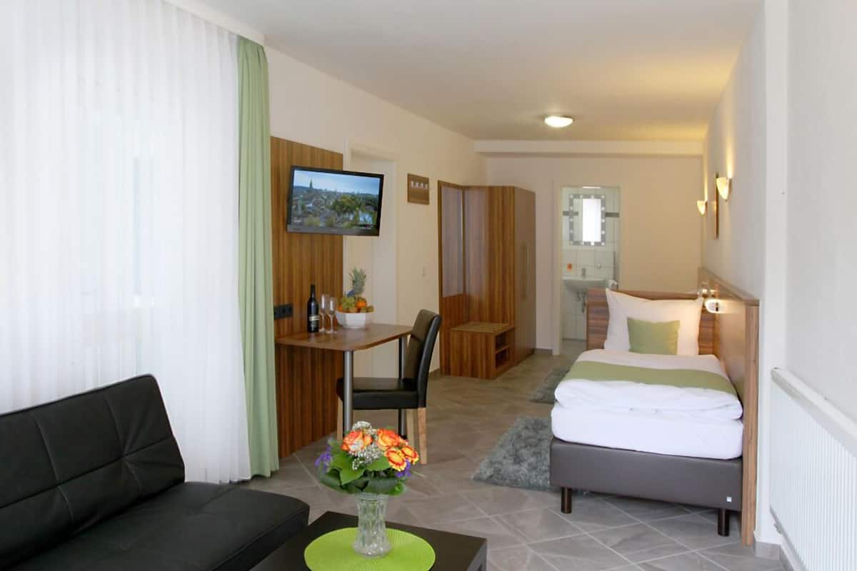 A hotel room with a bed, a sofa, a dining area with a table and chairs, a TV, and a bathroom with a mirror.