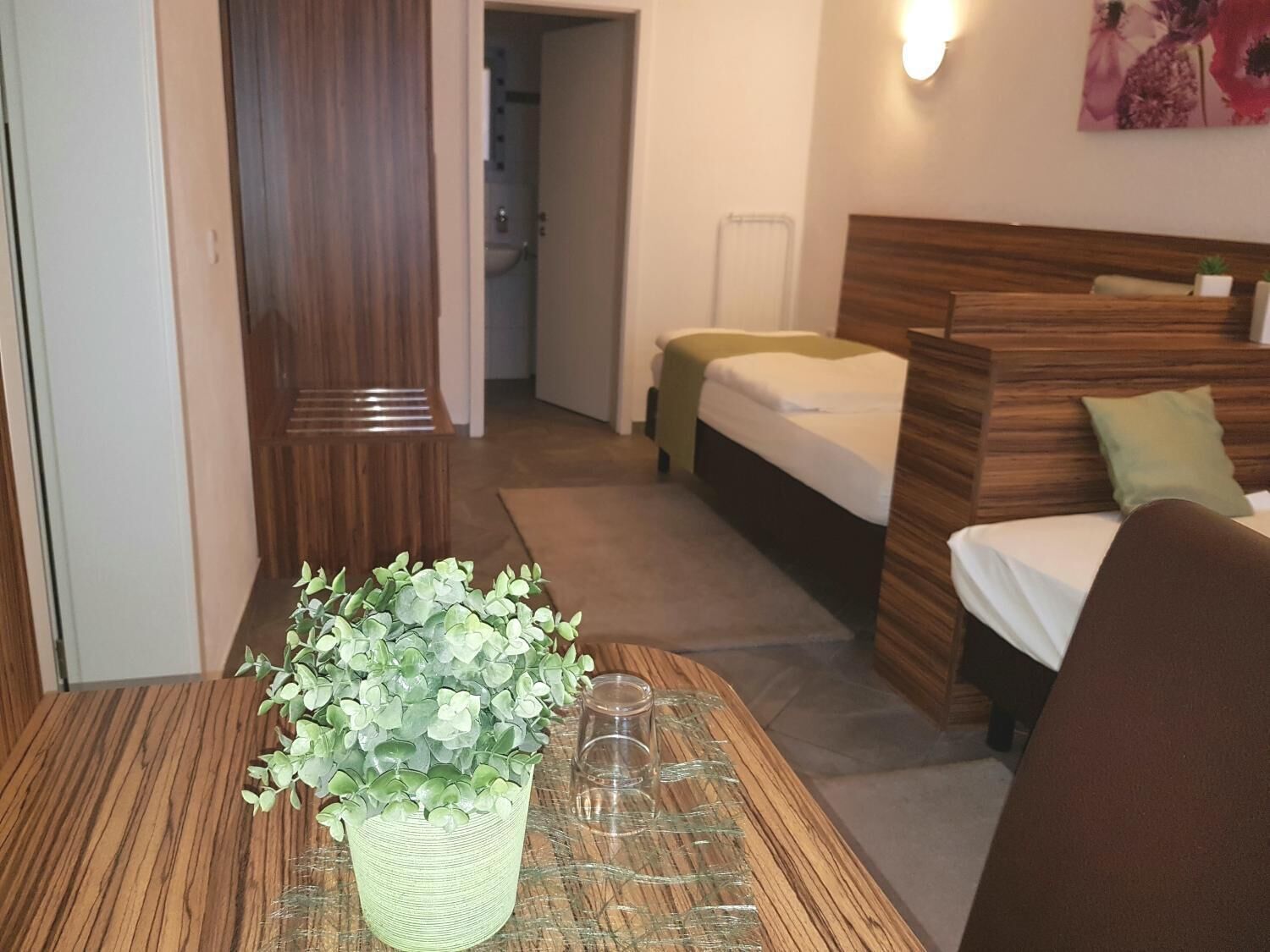 A hotel room with a wooden headboard, a bed with a green throw, a small table with a plant and glasses, and a wooden bench.