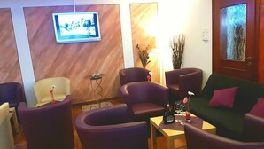 Uma sala de estar moderna com poltronas roxas, um sofá verde e uma pequena mesa com uma garrafa e copos. Uma televisão está fixada na parede e uma porta com painel de vidro jateado é visível.