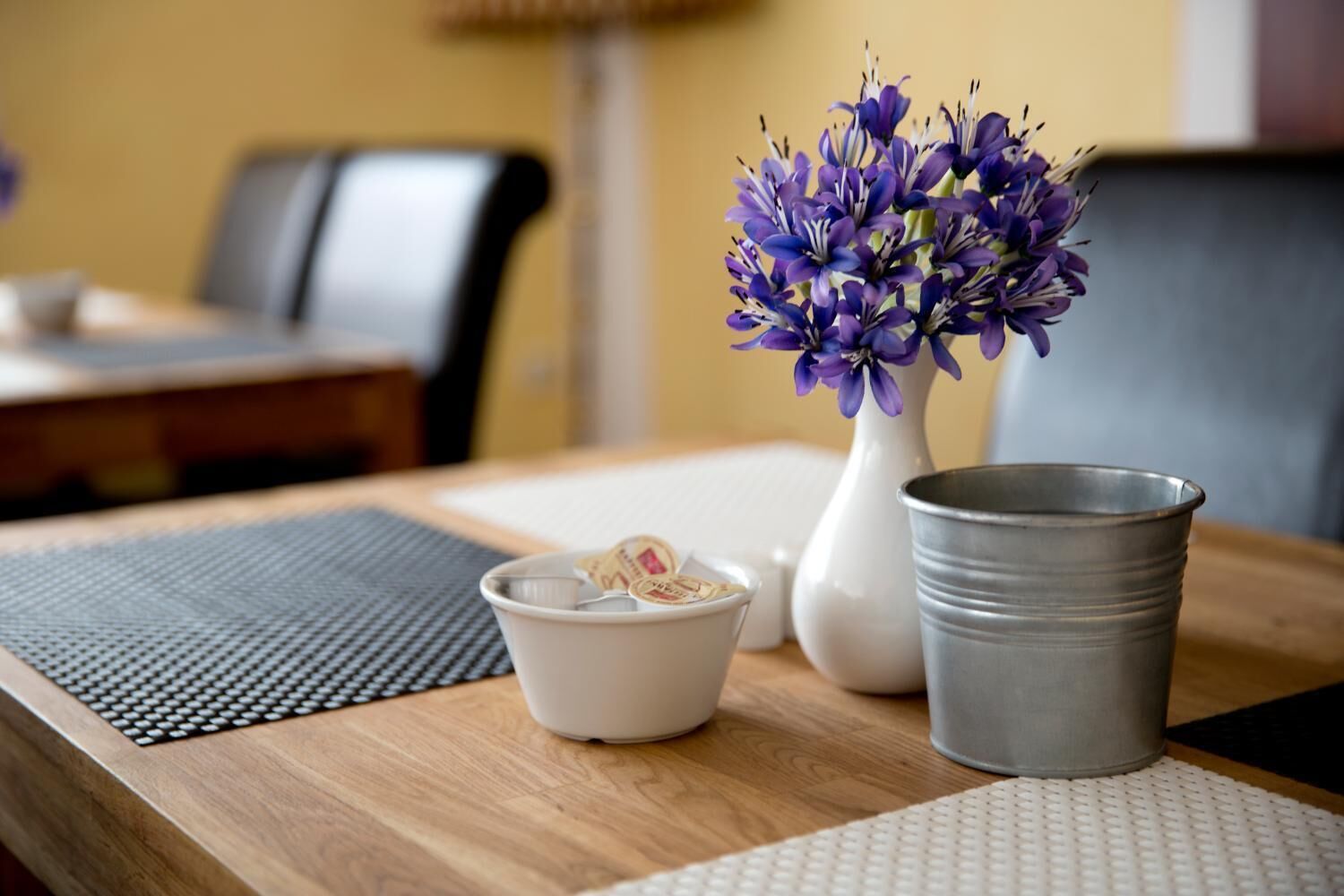 A wooden table with a white vase containing purple flowers, a small white bowl with tea bags, and a gray metal cup.