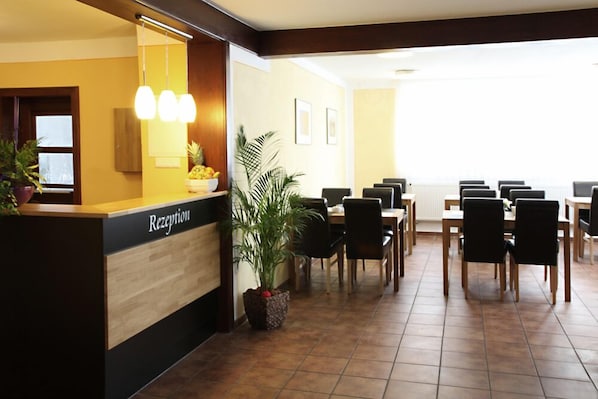 A reception area with a wooden reception desk, a large potted plant, and a dining area with multiple tables and chairs.