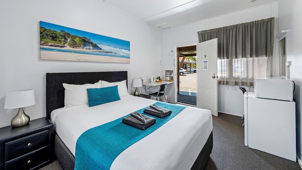 A hotel room with a bed, a desk, a refrigerator, and a painting of a beach. - Port Macquarie Motel (Port Macquarie)