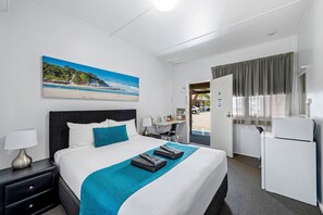 A hotel room with a bed, a desk, a refrigerator, and a painting of a beach. - Port Macquarie Motel (Port Macquarie)