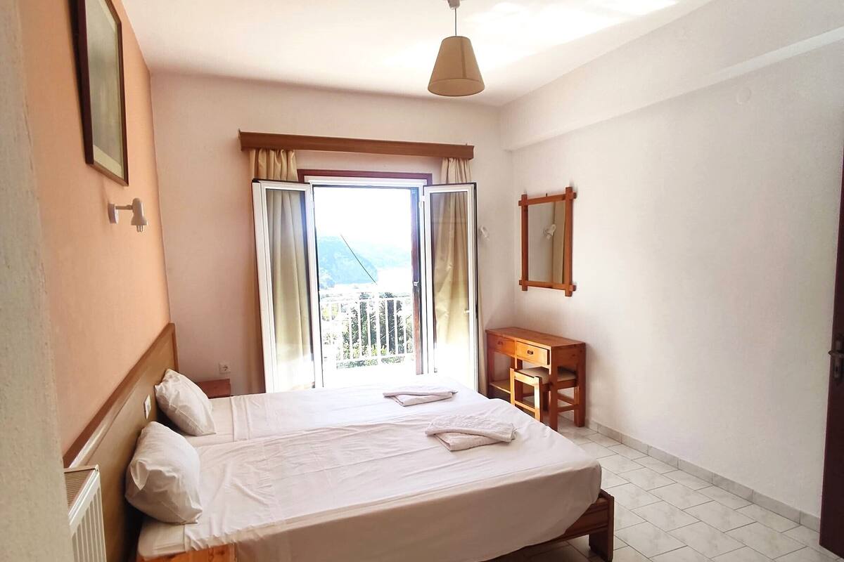A bedroom with a bed, a window with curtains, a small table, and a ceiling fan.