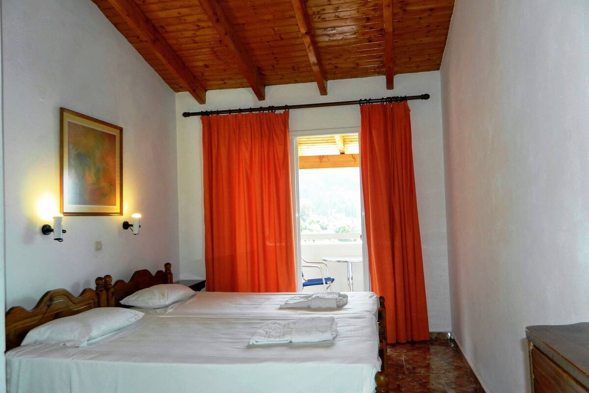 A bedroom with a wooden ceiling, a bed with white linens, orange curtains, and a painting on the wall.