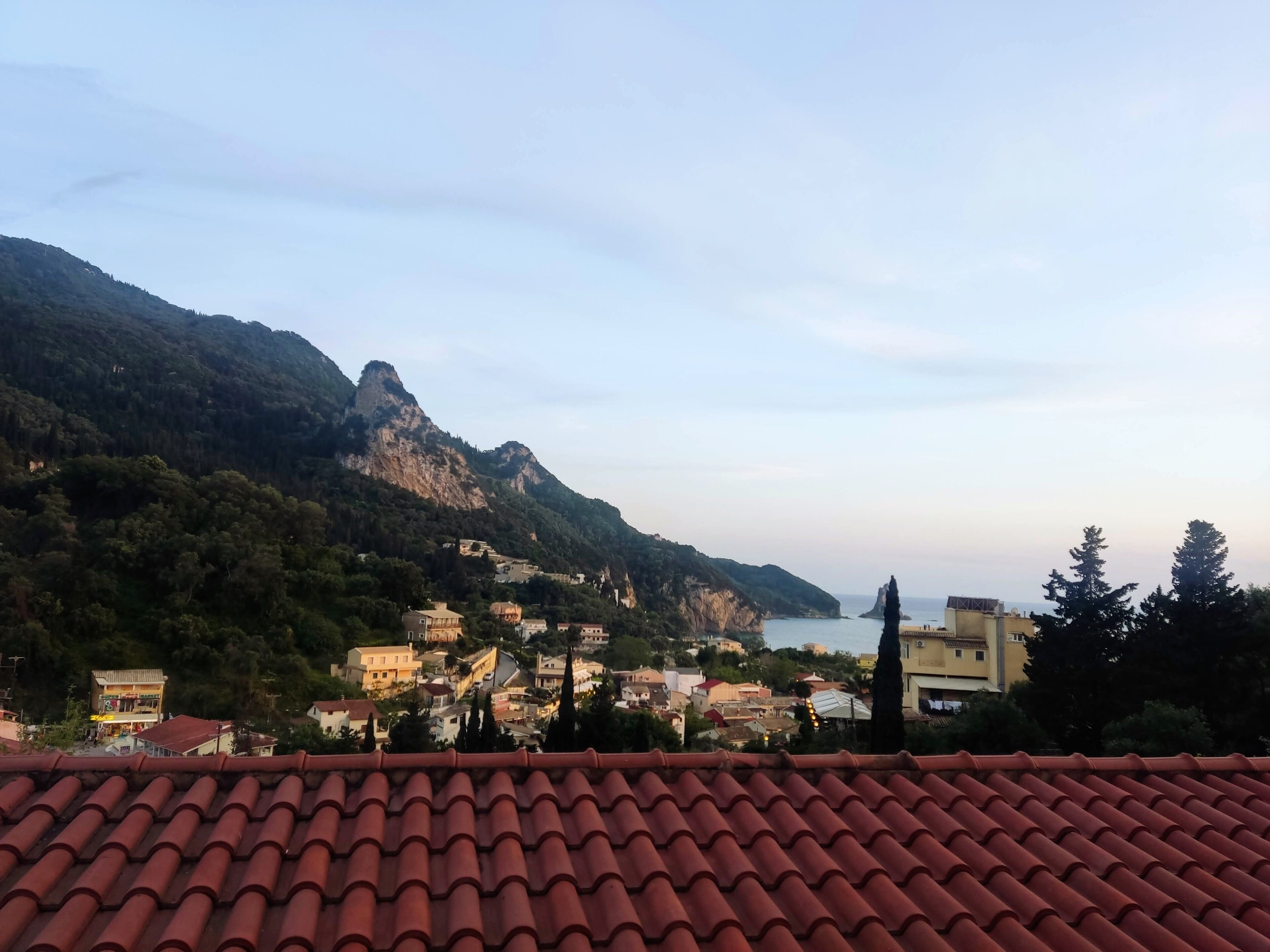 A coastal town with terracotta-tiled roofs, dense greenery, and a mountainous backdrop.