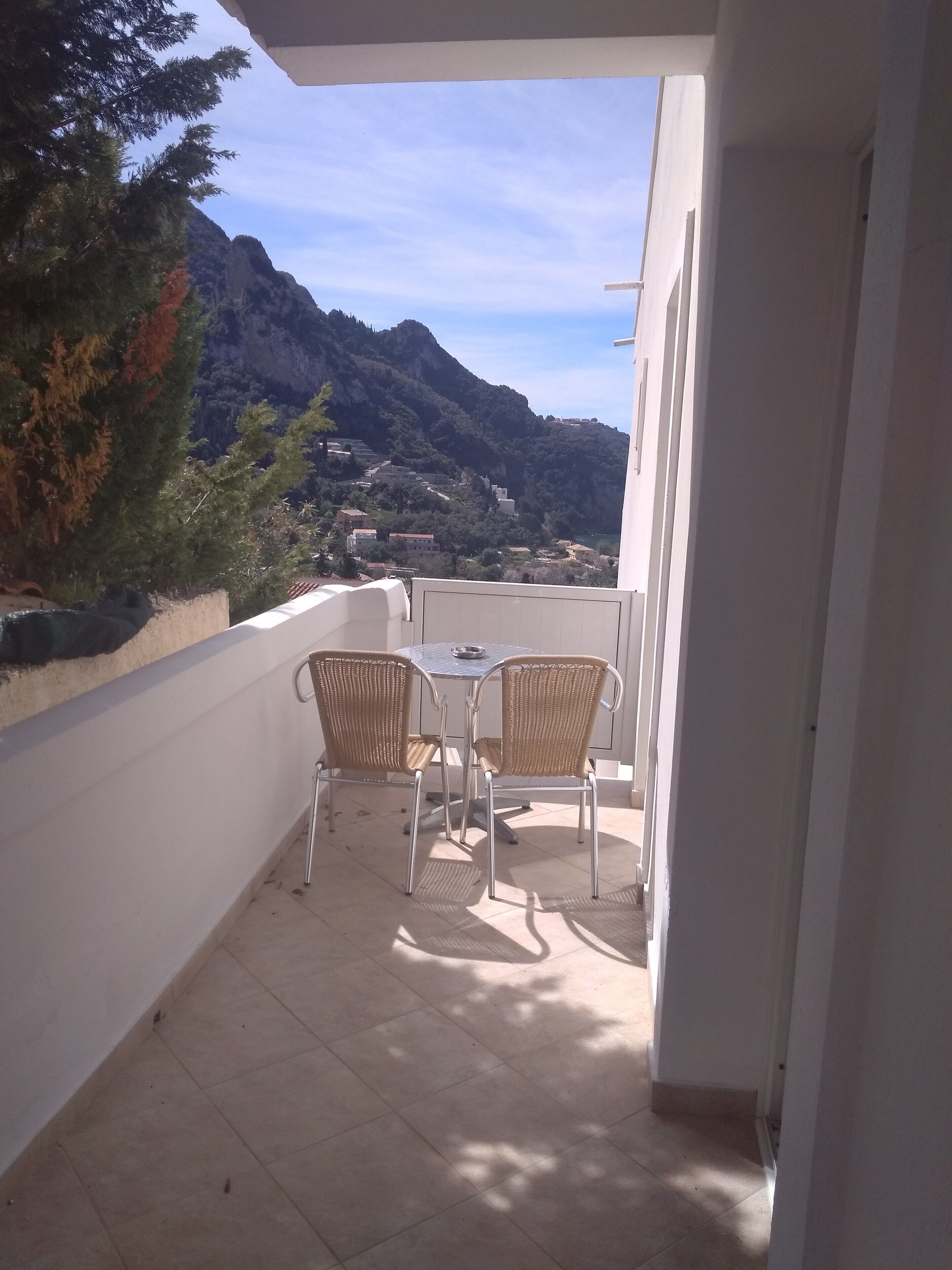 A balcony with two wicker chairs and a small table, overlooking a mountainous landscape with buildings.