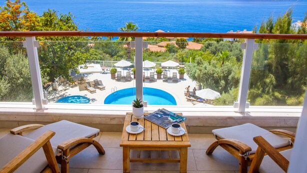 Un balcon avec une table en bois, deux fauteuils confortables et une vue sur une piscine et la mer.