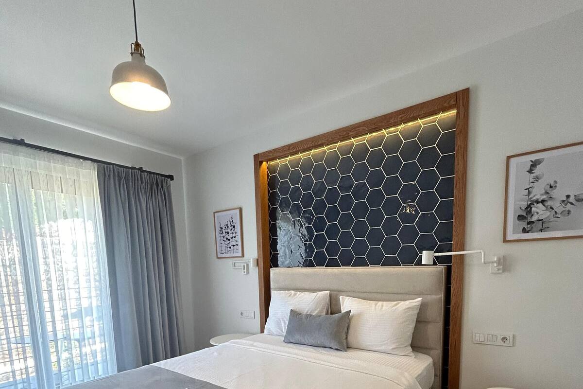 A modern bedroom with a large bed, a wooden headboard, a bedside table, and a view of the outdoors through a window with curtains.