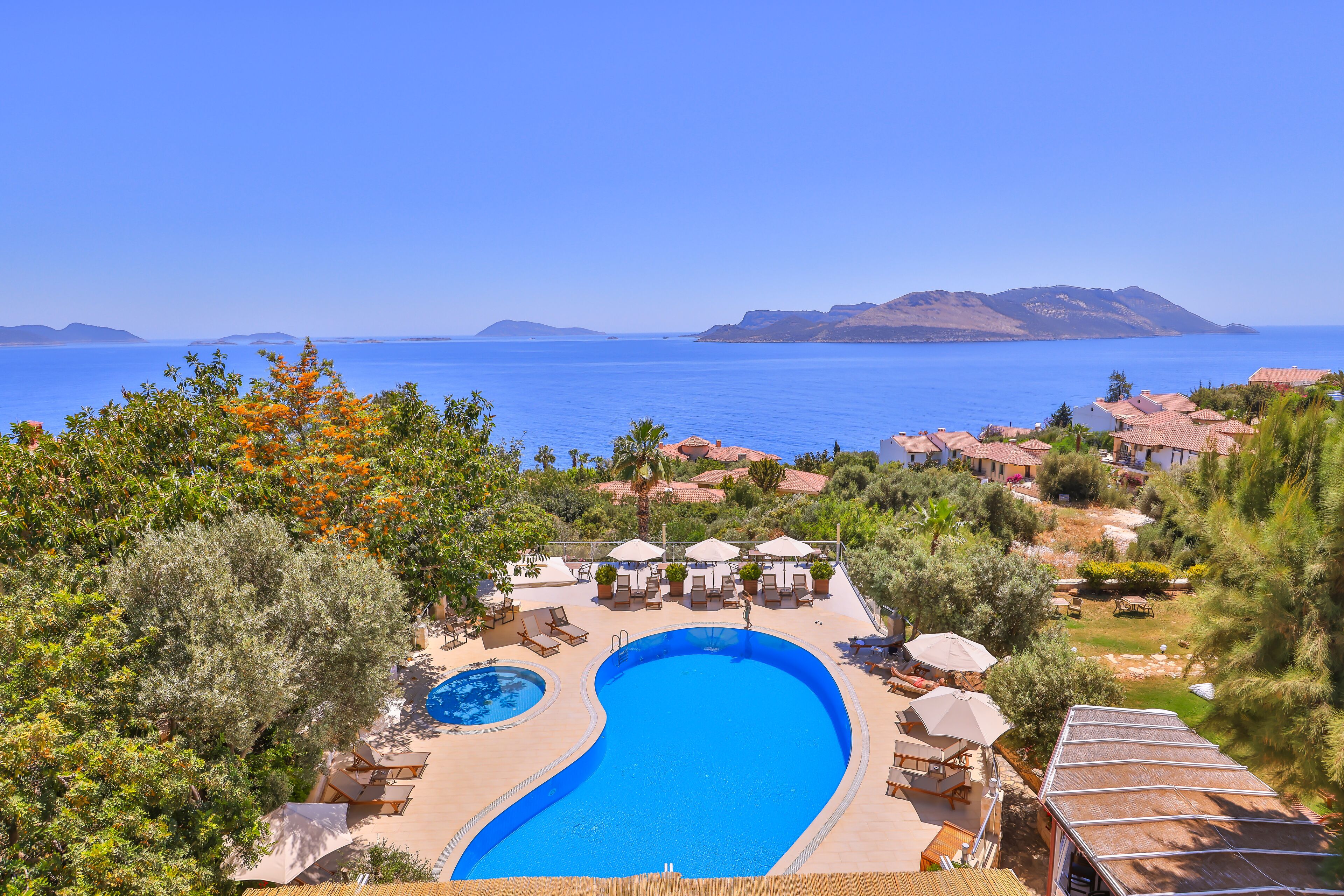 Un resort con una amplia piscina, sillas de descanso y sombrillas, rodeado de follaje y con vistas al mar azul y despejado.