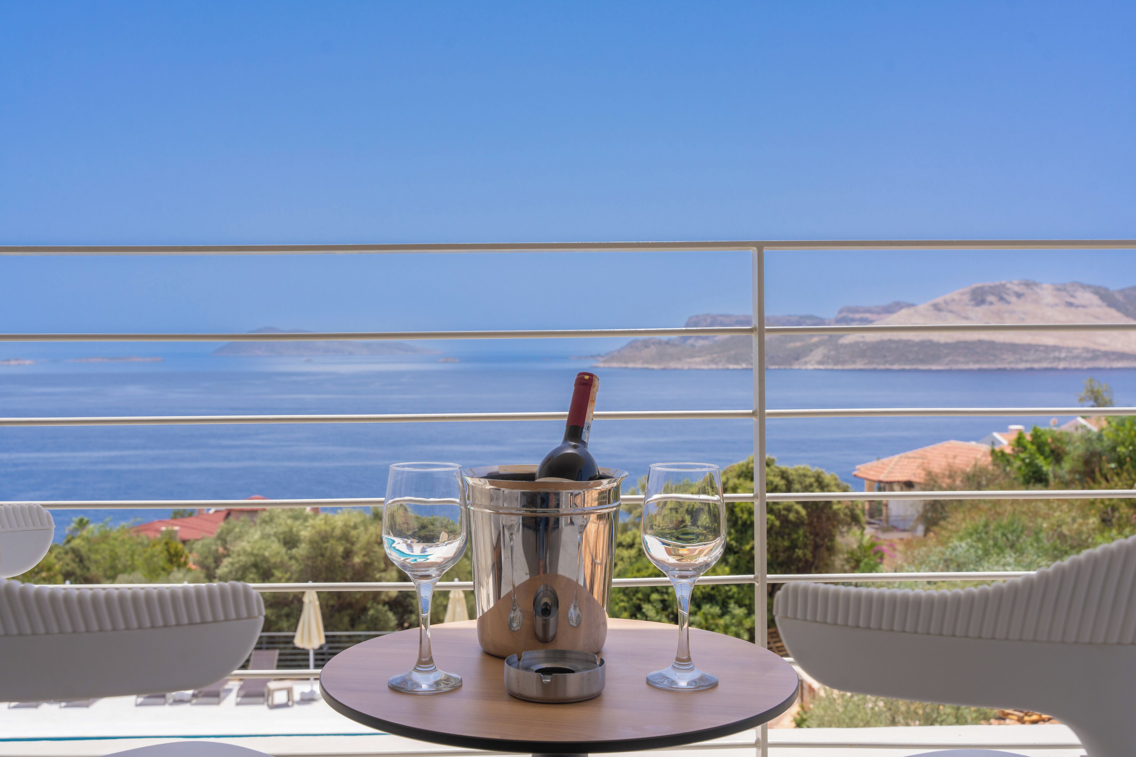A balcony with a table, two glasses, and a bottle of wine, overlooking a scenic view of the sea and hills.