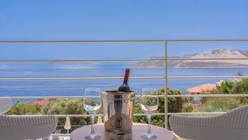 A balcony with a table, two glasses, and a bottle of wine, overlooking a scenic view of the sea and hills.