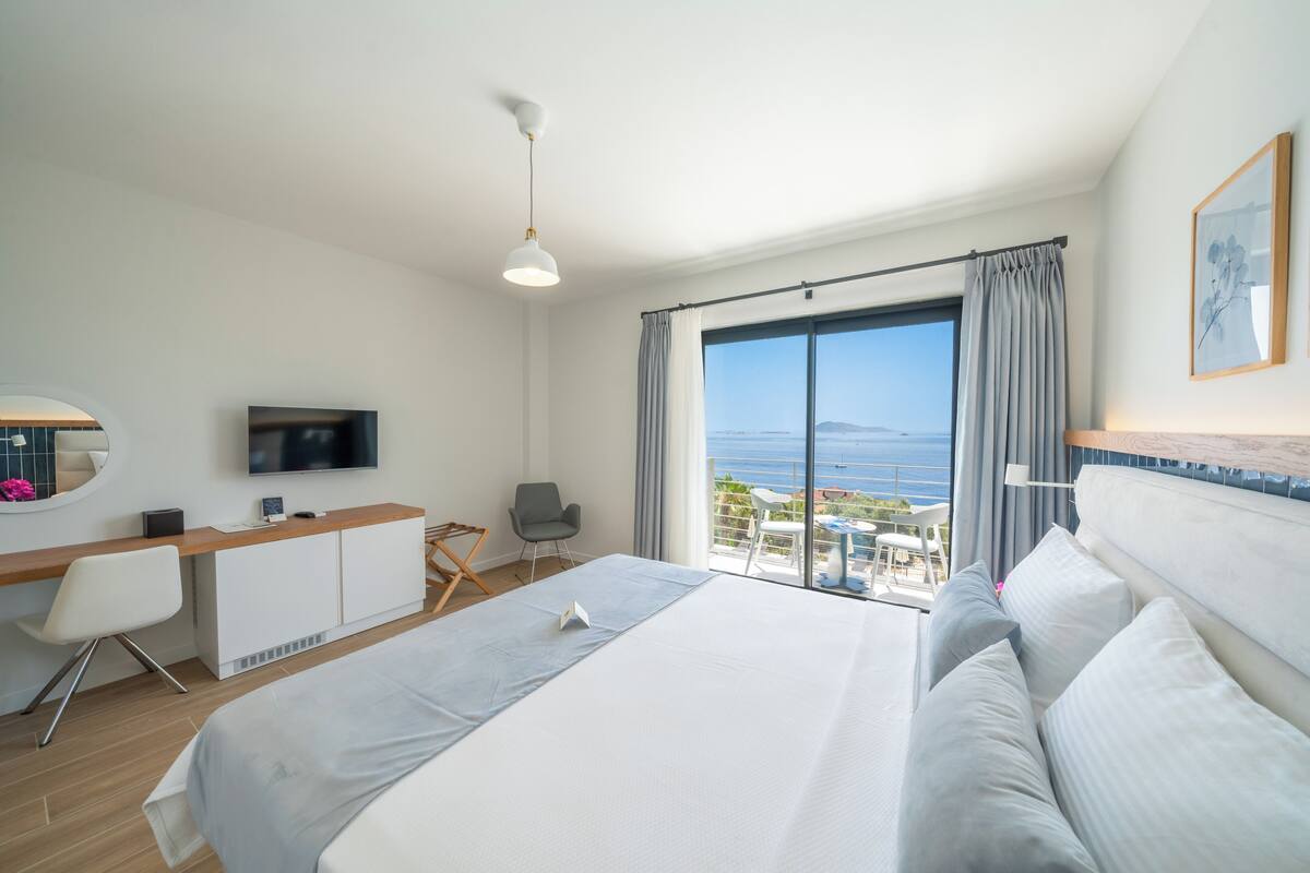 A hotel room with a large bed, a desk with a TV, a chair, and a view of the sea.