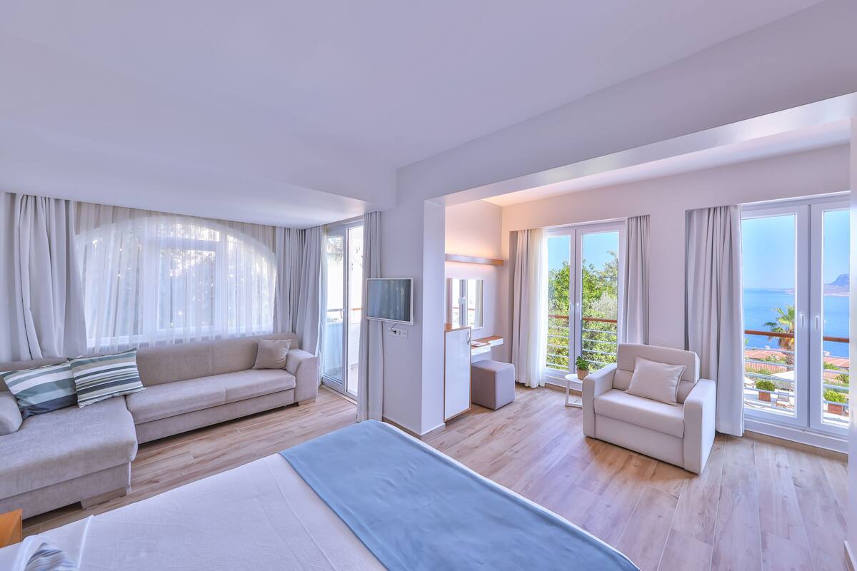 A modern hotel room with a large bed, a sofa, a chair, and a balcony with a view of the sea.