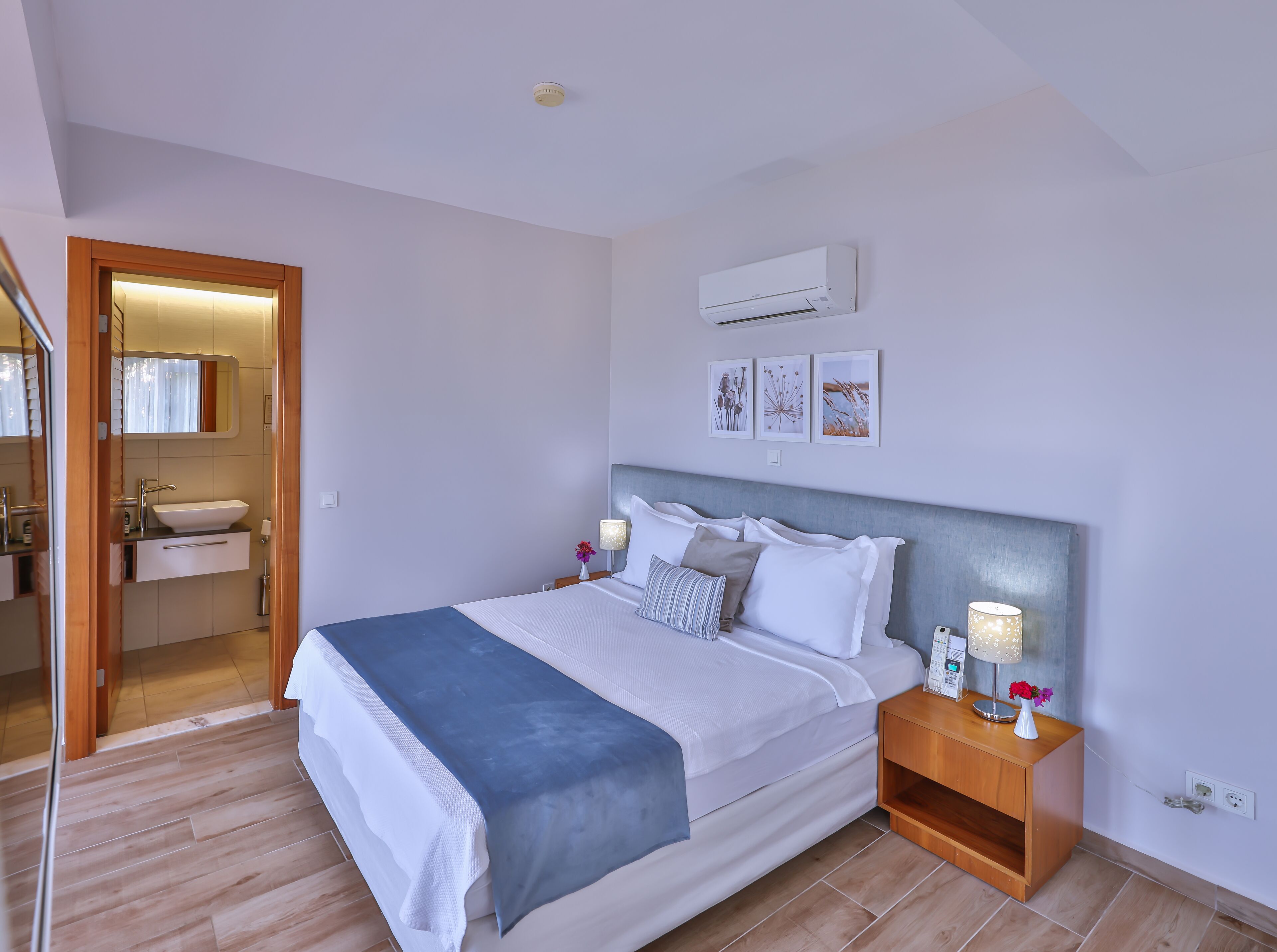 Une chambre moderne avec un lit, des tables de chevet, un climatiseur et une salle de bain visible à travers une porte ouverte.