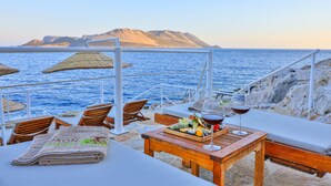 Une table avec un plateau de nourriture, des verres à vin, des chaises en bois et une vue sur la mer et les montagnes.