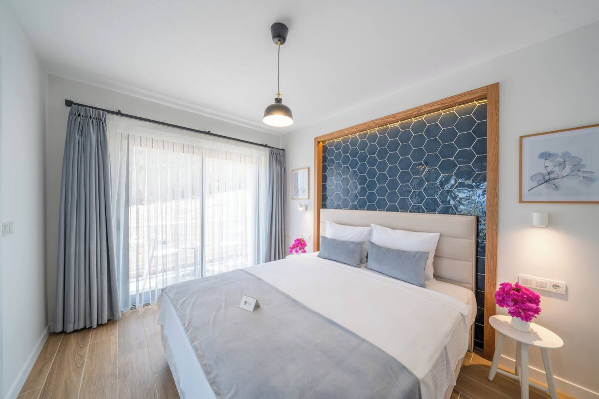 A modern bedroom with a large bed, a wooden headboard, a small bedside table with a vase of flowers, and a view of the outdoors through sheer curtains.
