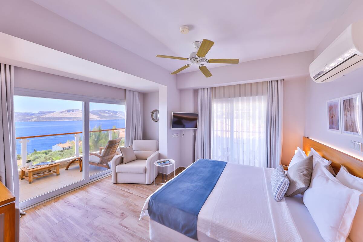 A hotel room with a bed, a TV, a chair, a small table, and a balcony with a view of the sea.