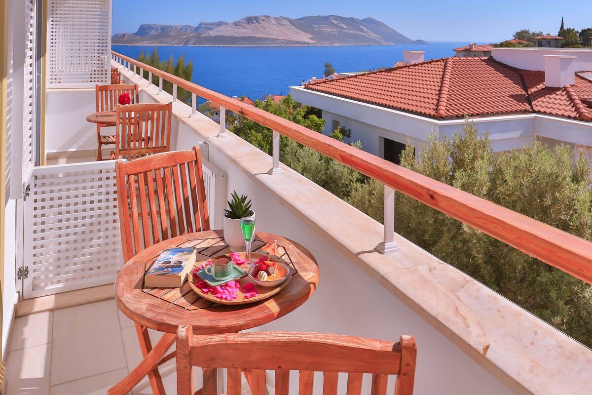A balcony with a table set for dining, overlooking a coastal view with mountains and a clear blue sky.